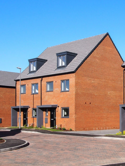 Two semi-detached red brick houses with a grey roof. A brick road is positioned in front of the house.