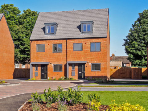 Two semi-detached red brick houses. A grass verge is positioned in front of the house with small shrubs.